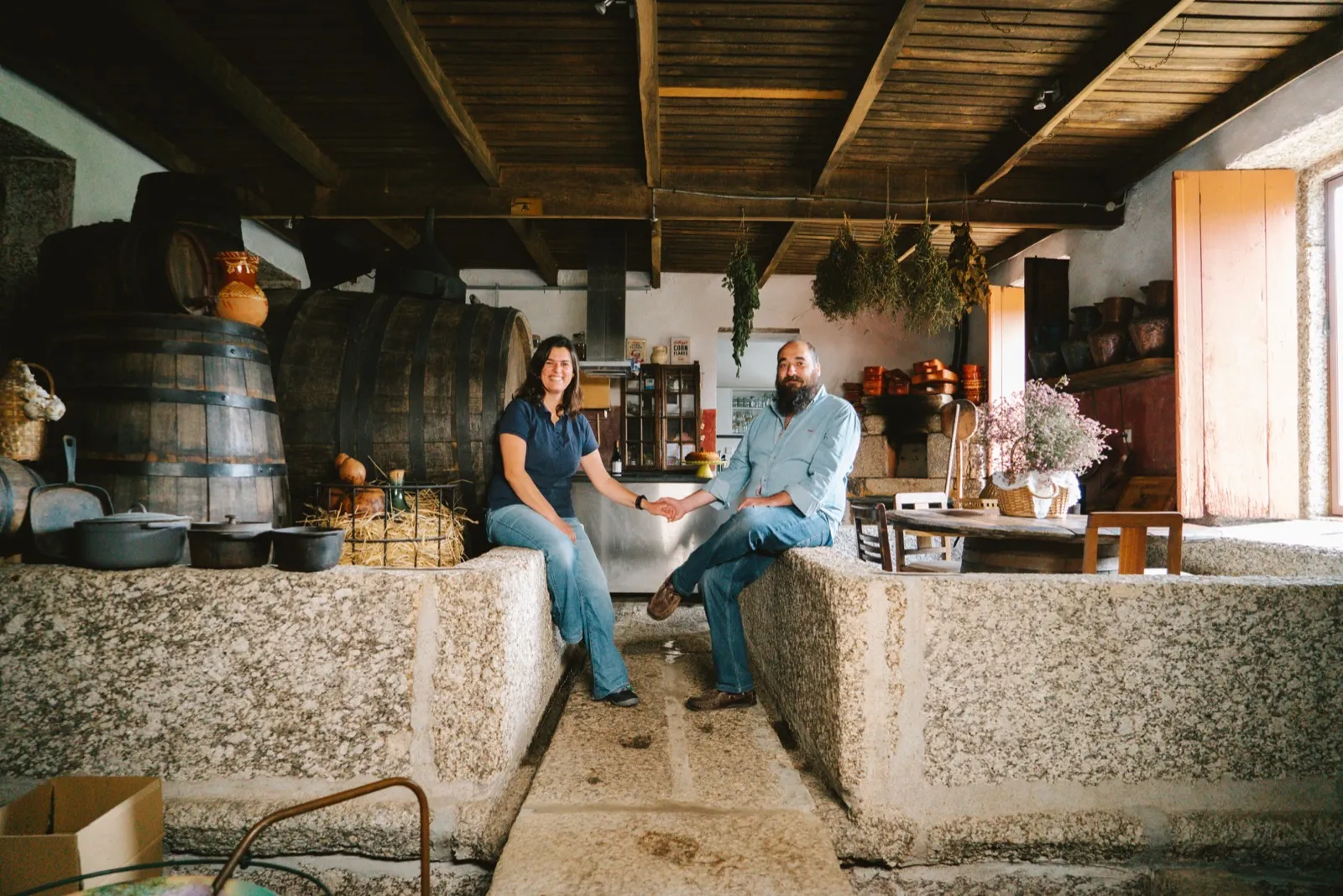 Teresa and Paulo holding hands in the old granite wine cellar, surrounded by barrels and drying herbs
